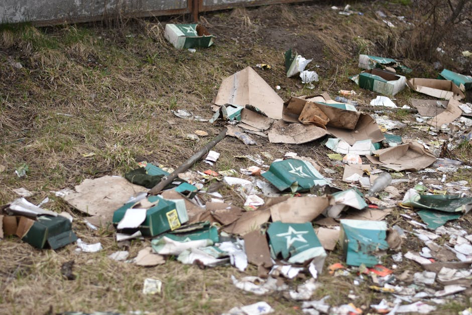 An outdoor scene showing scattered trash and rubbish on a patch of grass and soil, including torn cardboard boxes, some of which are green with white star logos, and various crumpled paper and plastic debris. The packaging appears damaged and disorganized, with pieces sprawled across the ground near a slight slope and against a wooden fence in the background. The environment looks neglected, möglicherweise in an area where waste has been dumped or left uncollected, highlighting the need for professional rubbish removal services such as those provided by House Clearance Chiswick to manage private disposal and prevent illegal dumping on residential or commercial sites.