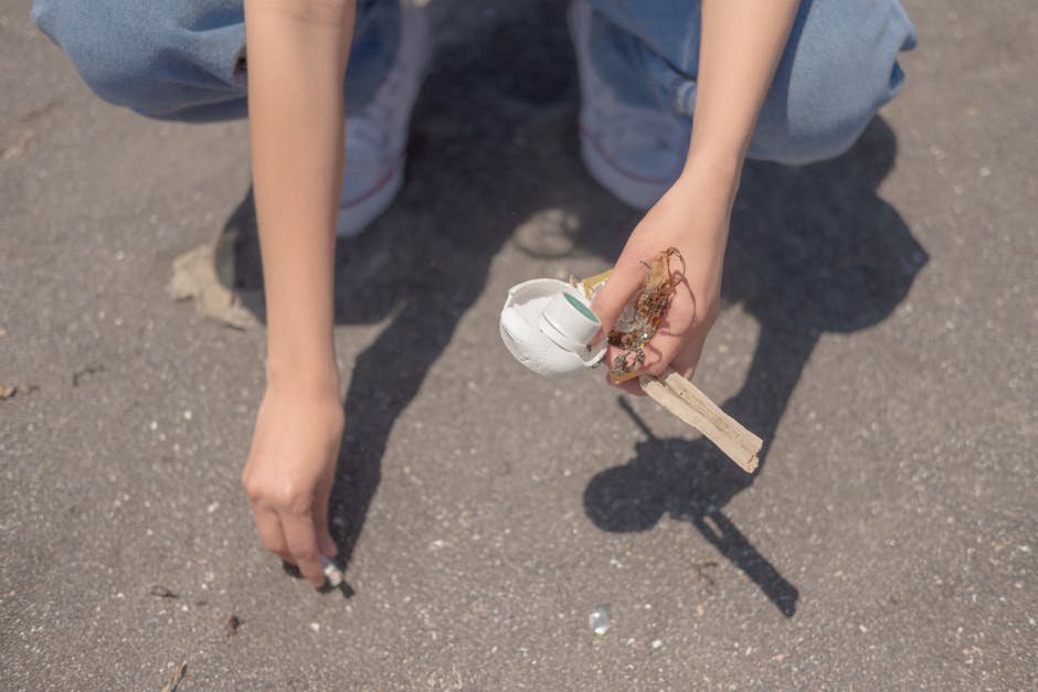 A person with fair skin, wearing light blue jeans and sneakers, crouches down on a paved surface outdoors during daytime. They are holding a small, broken ceramic cup with a rough, jagged edge, which appears to be white with a light blue interior, and bits of tangled string or twine attached to it. The person's right hand is reaching towards the ground, while their left hand loosely grasps the broken piece. The scene is clearly lit by natural light, casting shadows of the person's arms and the broken ceramic piece onto the ground. Scattered around are small fragments of debris and dirt, suggesting the area is used for informal rubbish collection or site clearance. The background shows a section of the paved outdoor environment, emphasizing a setting where trash or discarded objects are being gathered or removed, aligning with services that handle on-site waste clearance. House Clearance Chiswick offers such independent waste handling solutions, exemplified by the discarded ceramic piece in the person's possession.