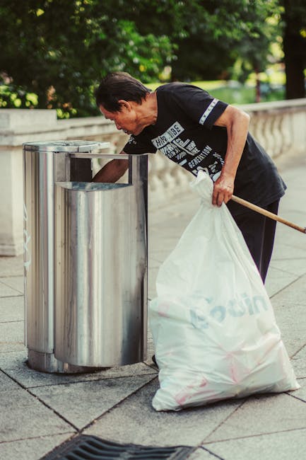 A middle-aged man with dark hair, dressed in a black T-shirt with white printed text on the front and dark trousers, is seen using a wooden stick to guide a large white plastic bag full of collected waste into a metallic litter bin. The bin has a cylindrical shape with a shiny, reflective surface and an open top, positioned on a paved sidewalk. In the background, there is a low stone wall and lush green foliage, indicating an outdoor setting in a park or residential area. The man is bent slightly forward, focused on his task of waste collection, which is part of an outdoor rubbish removal activity. The environment appears clean and well-maintained, with natural daylight illuminating the scene, highlighting the textures of the metal bin, the plastic bag, and the surrounding greenery. This scene reflects a community effort in local waste management or private rubbish handling, associated with services such as House Clearance Chiswick.