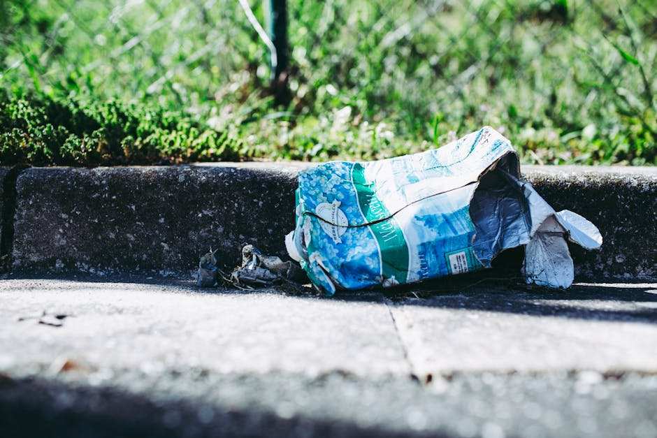 A crumpled plastic bag made of thin, flexible material with a glossy surface lies discarded on a concrete pavement, partially resting against the edge of a curb. The bag features a colorful printed design with shades of blue, green, and white, including images of landscapes and text, indicating it was once used for shopping or packaging. The pavement appears rough and weathered, with small cracks and dirt visible nearby. The curb, constructed from dark, textured concrete, separates the sidewalk from a grassy area beyond, which contains lush green grass and sparse weeds, illuminated by natural daylight. The scene suggests a typical urban or suburban street environment where waste has been improperly disposed of outside designated rubbish collection points. This visual context supports the services of companies like House Clearance Chiswick that handle private waste removal and on-site clearance, helping to maintain environmental standards and comply with local rubbish regulations.
