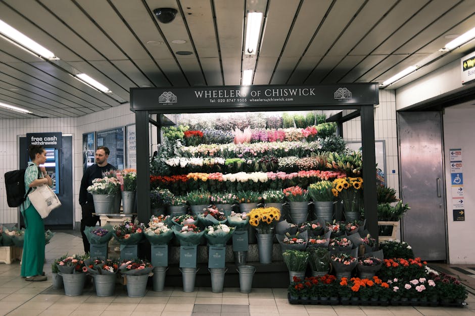 Inside a well-lit underground station with a white tiled ceiling fitted with long fluorescent light fixtures, there is a flower kiosk named 'Wheeler of Chiswick' that displays an extensive collection of potted flowering plants arranged on multi-tiered shelves and in buckets. The flowers include a variety of colourful blooms such as white, pink, yellow, red, and purple, with some plants featuring lush green foliage. To the left, a woman with dark hair tied back, dressed in teal trousers, a patterned top, and carrying a black backpack and a white tote bag, is engaged in conversation with a man wearing a dark sweatshirt, who is standing behind the flower display. The background features a cash machine marked 'free cash withdrawal' on the wall, with informational notices nearby, and doors leading to other parts of the station. The scene conveys a busy urban setting where commuters might purchase flowers en route, highlighting the station's role as an accessible spot for informal or private waste and product transaction activities, and exemplifying the importance of maintaining a clean environment as per local rubbish regulations.