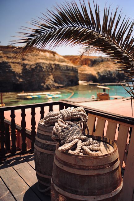 Two wooden barrels with metal bands, positioned on a wooden deck, each containing thick coiled ropes with textured, weathered surfaces. The barrels are placed close together, with the ropes loosely arranged on top, showing areas of fraying and roughness. Behind the barrels, there is a railing made of dark wood balusters, and beyond that, a scenic coastal view features clear turquoise water, rocky cliffs, and a sandy shoreline. A large palm frond arches over the scene from the upper left corner, partially shading the barrels and ropes, contributing to a tropical seaside atmosphere. The lighting indicates bright, natural sunlight, casting soft shadows on the deck. This setting suggests outdoor maritime or coastal storage, typical in destinations where informal or alternative waste handling may occur, aligning subtly with the theme of external rubbish or equipment removal, as managed by local services like House Clearance Chiswick.