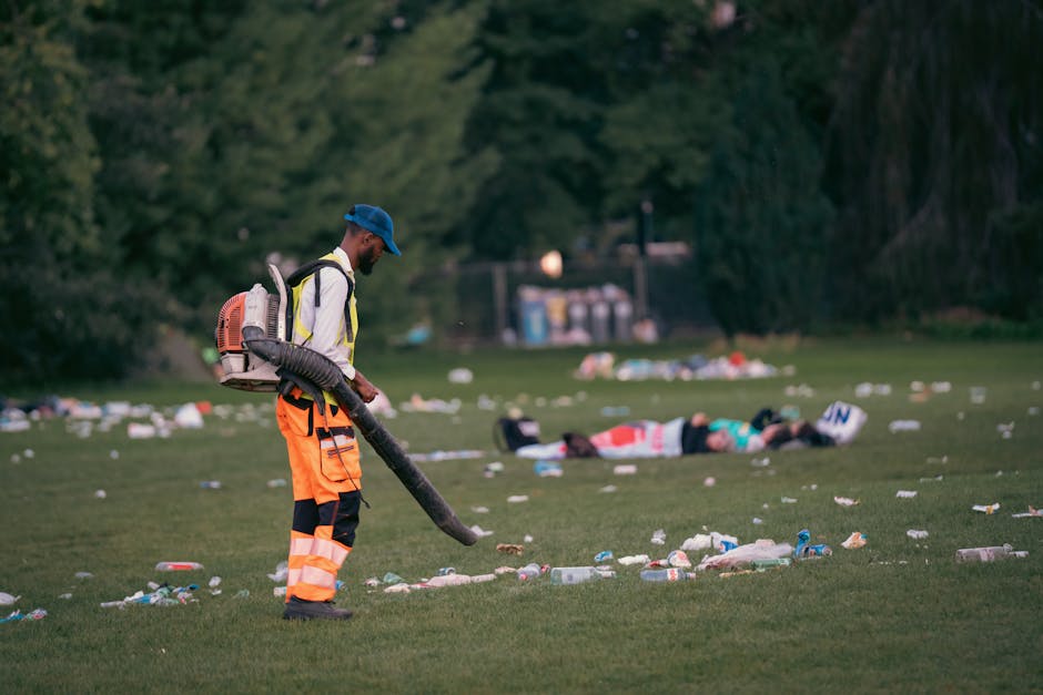A male worker dressed in high-visibility orange trousers with reflective strips, a yellow jacket, and a blue cap is standing on a grassy area, using a leaf blower or similar equipment to clear debris. The grass field around him is scattered with numerous plastic bottles, cups, and other rubbish, indicating a recent littering incident or incomplete cleanup. In the background, there are two people lying on the grass, possibly resting or affected by the cleanliness of the area. Behind them, tall trees and a row of waste bins are visible, situated near a fence, suggesting a park or recreational space. The natural lighting implies early evening or late afternoon. The scene highlights the importance of waste clearance in outdoor environments, aligning with private rubbish removal services such as those provided by House Clearance Chiswick, who offer solutions for managing accumulated rubbish in public and private spaces to support environmental cleanliness and hygiene.
