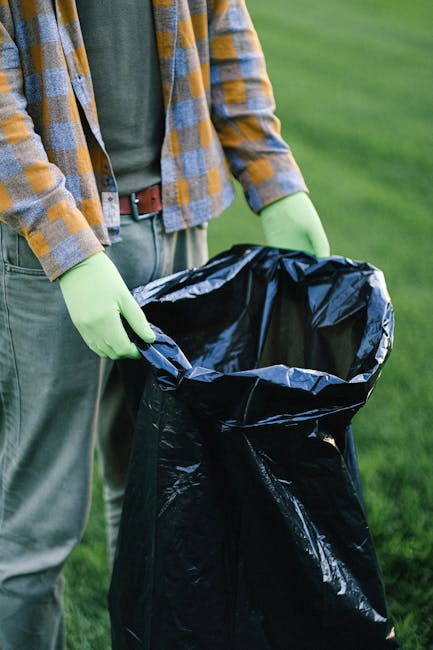A person is standing outdoors on a grassy area, wearing a casual plaid shirt in shades of yellow, gray, and white, along with gray trousers and bright green gloves. They are holding a black plastic rubbish bag open, which appears to be partially filled, ready for waste collection. The setting suggests an activity such as rubbish or waste collection, possibly part of a nearby garden or outdoor clearance. The background shows a well-maintained green lawn, with no other objects or structures visible. The image emphasizes the preparation for independent waste disposal or rubbish removal, typical of private or non-authority managed services. The natural lighting highlights the textures of the person's clothing, the glossy surface of the rubbish bag, and the vibrant greenery surrounding them, contributing to a clear, professional, and neutral visual representation of outdoor rubbish collection activities, aligned with services such as those offered by House Clearance Chiswick.