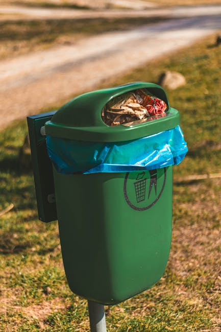 A close-up view of a green outdoor rubbish bin situated on a grassy area with a dirt pathway in the background. The bin has a rounded, dome-shaped lid partially transparent, revealing mixed waste including foil wrappers and plastic packaging inside. A blue plastic liner is visible around the opening, secured beneath the lid. The bin features a black icon of a recycling bin with a person disposal symbol on its front. The environment appears natural with soft sunlight illuminating the scene, and the bin is positioned on a metal post anchoring it to the ground. This setting exemplifies an independent waste disposal station, typical of private rubbish collection services that handle waste beyond local authority provisions, aligning subtly with themes of on-site clearance and alternative rubbish disposal services like those offered by House Clearance Chiswick.
