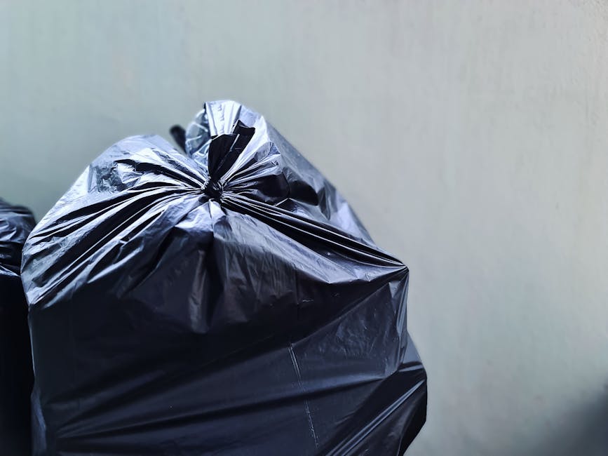 Close-up view of several large black plastic rubbish bags with shiny, crinkled textures, tightly tied at the tops, and stacked against a plain, light-colored wall. The bags are placed on a flat surface, possibly a driveway or a pavement, and are intended for waste collection or disposal. The background is minimal, emphasizing the focus on the waste bags, which are part of an informal rubbish removal process, potentially handled by a private service like House Clearance Chiswick. The lighting is neutral and diffuse, highlighting the reflective surfaces of the plastic material and the contours created by the gathered knots at the tops of the bags, indicating a recent accumulation of household or garden waste meant for an alternative waste management solution.